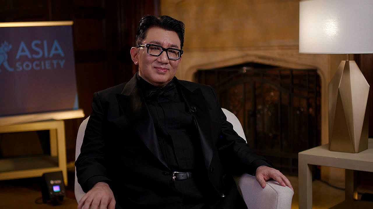 Homem de terno preto e óculos participa de entrevista em ambiente interno com painel da Asia Society ao fundo