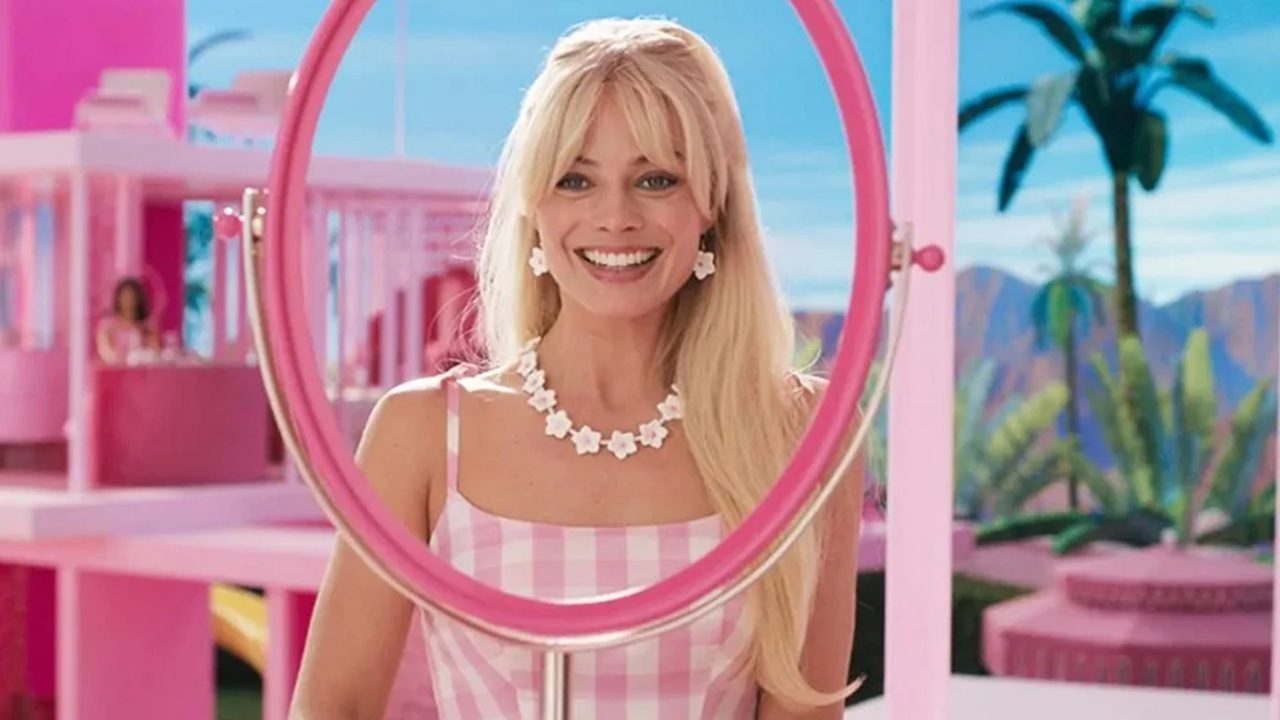 Mulher loira sorri em frente a um espelho circular rosa em um cenário vibrante e inteiramente cor-de-rosa, com arquitetura estilizada e palmeiras ao fundo. Ela usa vestido xadrez rosa e branco, além de colar e brincos de flores brancas, transmitindo estética lúdica e decorativa