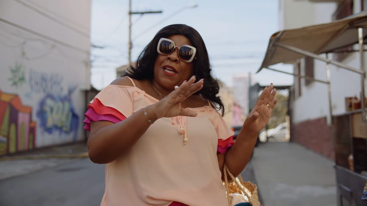 A imagem mostra uma mulher em uma rua urbana, usando óculos de sol grandes e blusa em tons de pêssego e rosa com babados nas mangas. Ela gesticula com as mãos, aparentando estar em uma conversa animada. Ao fundo, há muros grafitados e barracas de rua, compondo um cenário descontraído e cotidiano