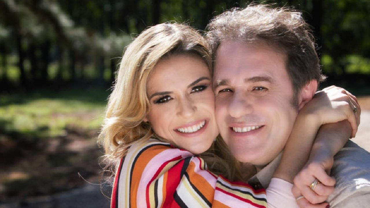 Casal sorri abraçado em ambiente ao ar livre, com fundo de árvores e luz natural; a mulher veste blusa listrada colorida e ambos aparentam felicidade e carinho