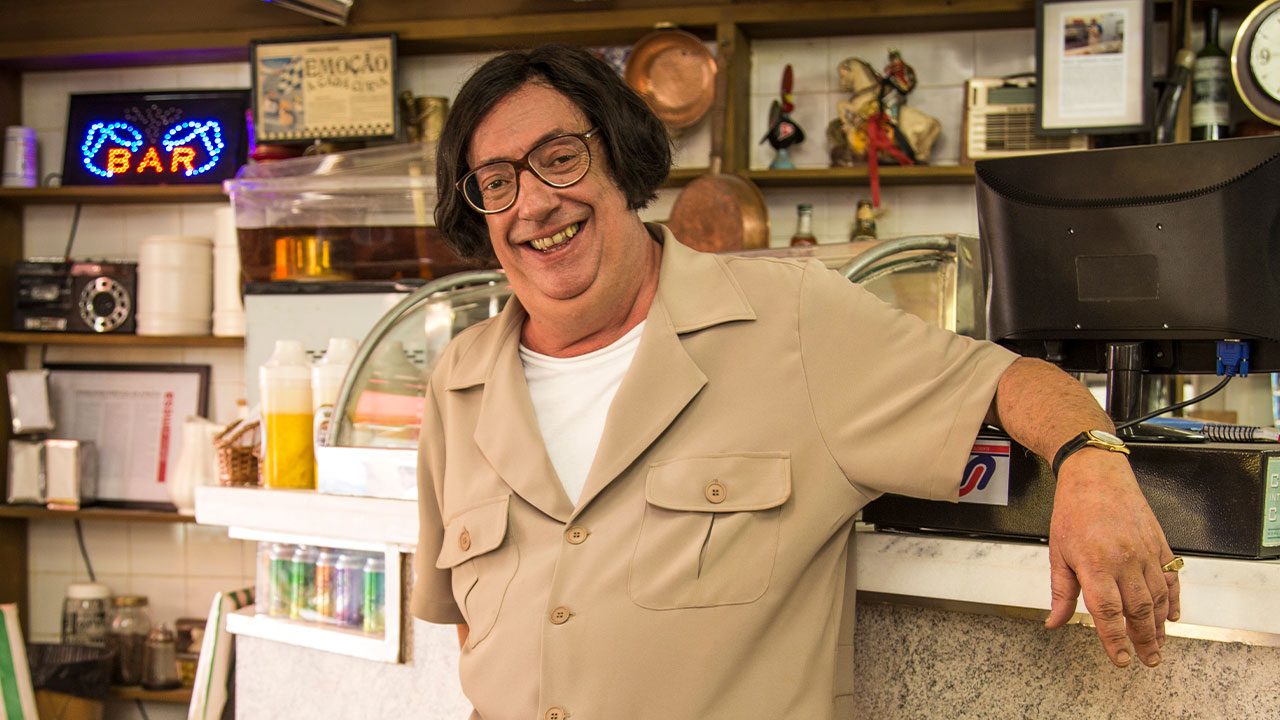 Marcos Oliveira caracterizado como Beiçola encostado no balcão da pastelaria em A Grande Família com sorriso no rosto