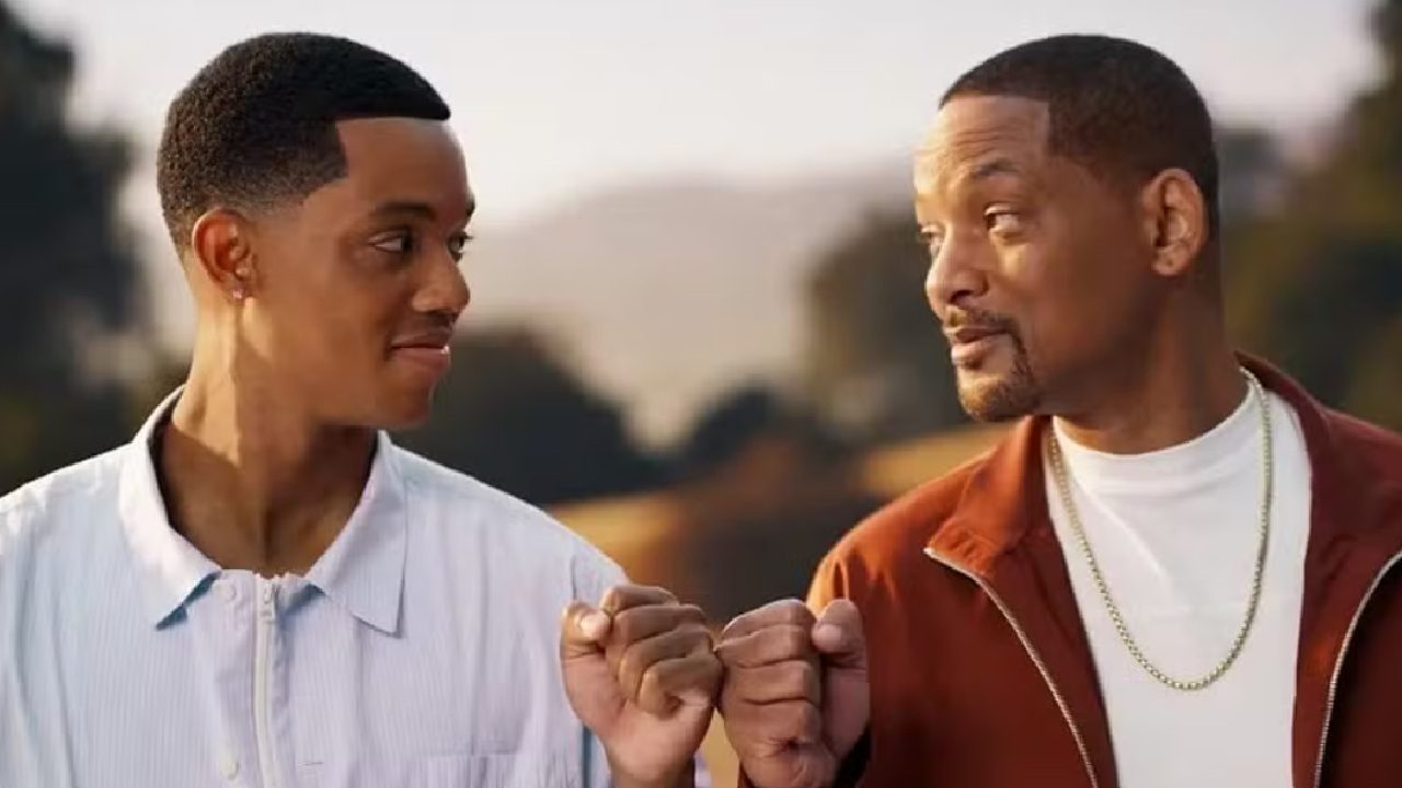 Dois homens se encaram com expressão amistosa enquanto encostam os punhos em cumprimento; ambos sorriem levemente ao ar livre, com fundo desfocado de árvores ao entardecer