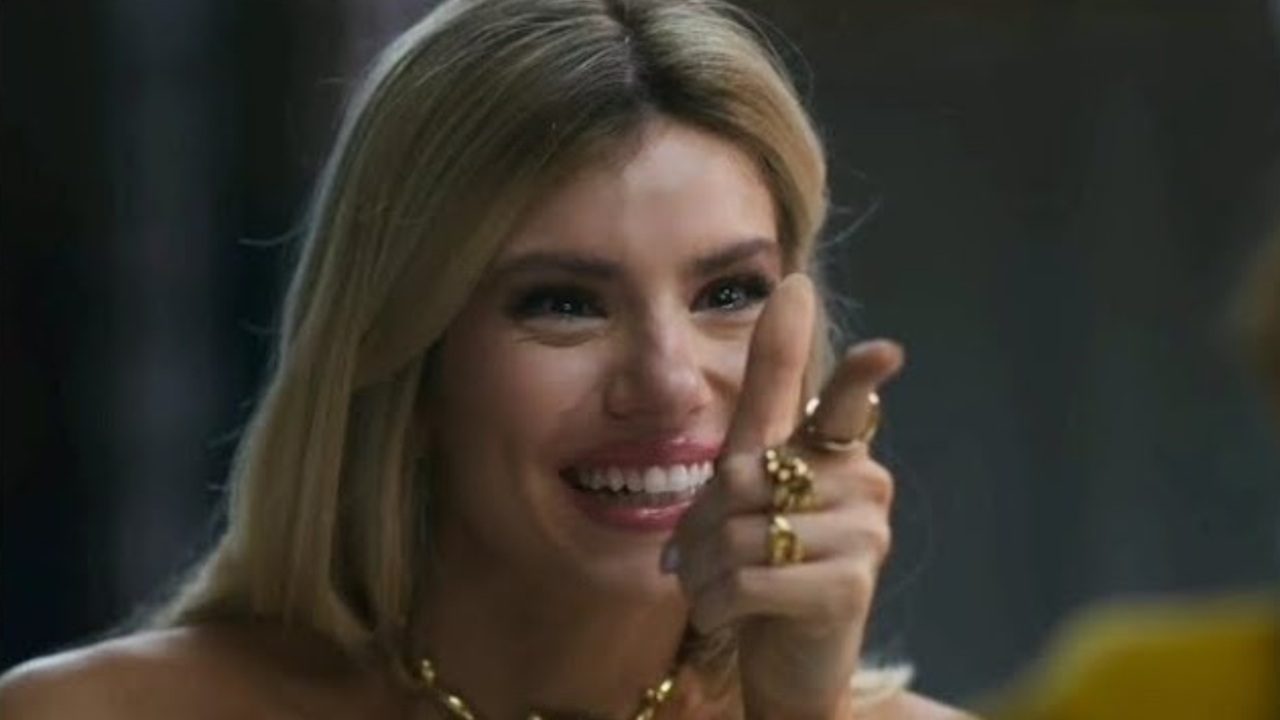 Mulher sorridente, com cabelos loiros soltos e acessórios dourados, aponta para frente de forma descontraída, exibindo anéis e colar enquanto olha para alguém fora do quadro