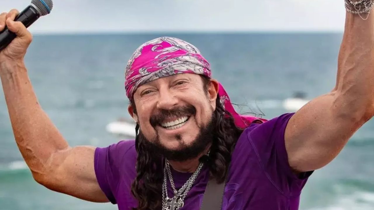 A imagem mostra um homem sorridente ao ar livre, segurando um microfone e vestindo uma camiseta roxa. Ele usa um lenço rosa estampado na cabeça, colares prateados e tem barba e cabelo comprido. Ao fundo, é possível ver o mar, sugerindo que ele está em uma apresentação ou evento próximo à praia