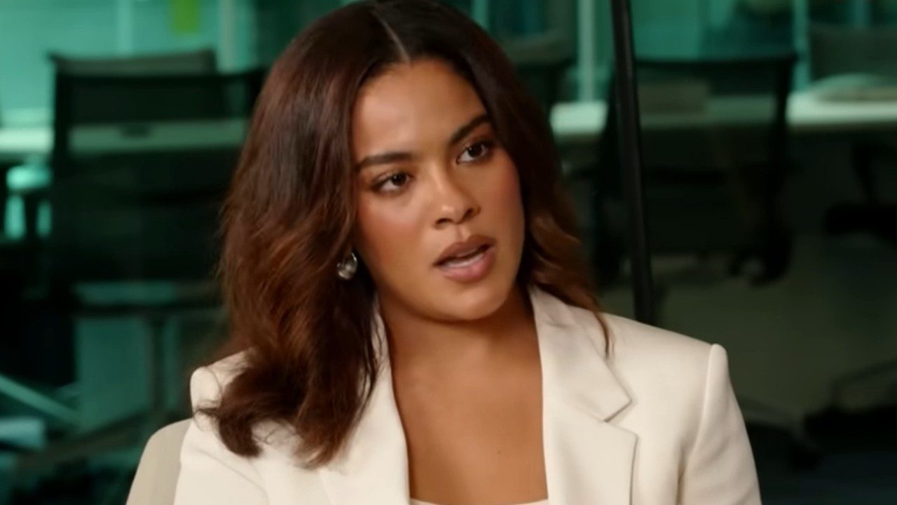 Bella Campos com blazer claro e cabelo solto fala durante entrevista em ambiente interno com cadeiras ao fundo