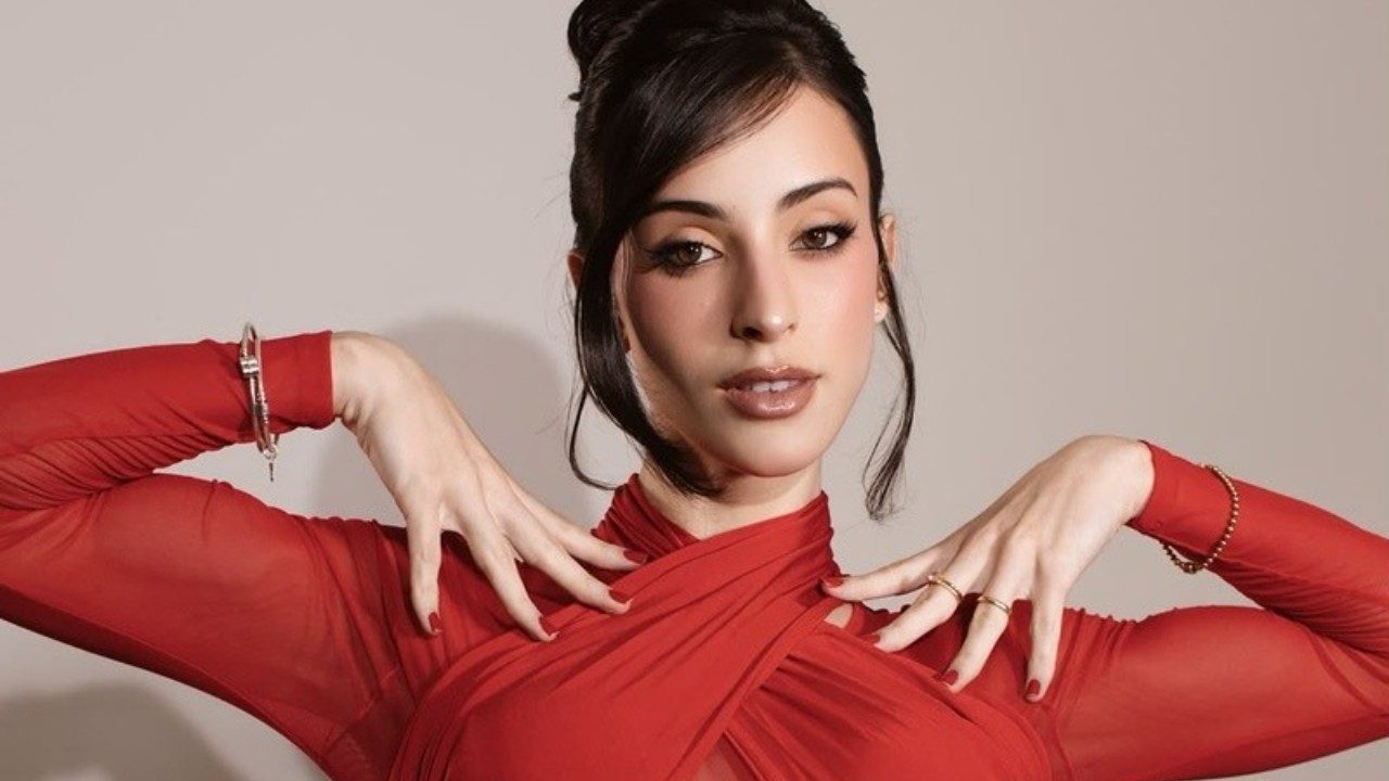 Bella Longuinho posa com vestido vermelho de mangas transparentes e mãos junto ao rosto diante de fundo neutro