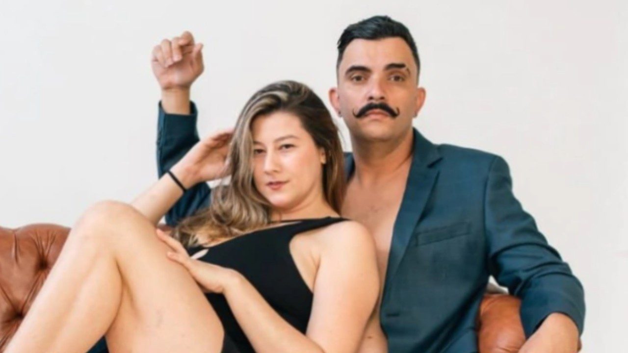 Bella Mantovani, de maiô preto, e Vagner Macedo, de terno cinza e bigode marcante, posam sentados em um sofá de couro marrom com fundo branco neutro