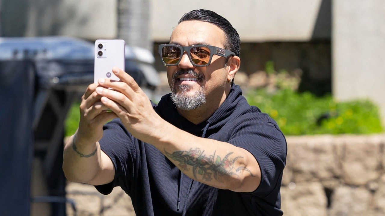 Cantor Belo usa óculos escuros e segura celular para foto ao ar livre nos Estúdios Globo com tatuagens visíveis no braço