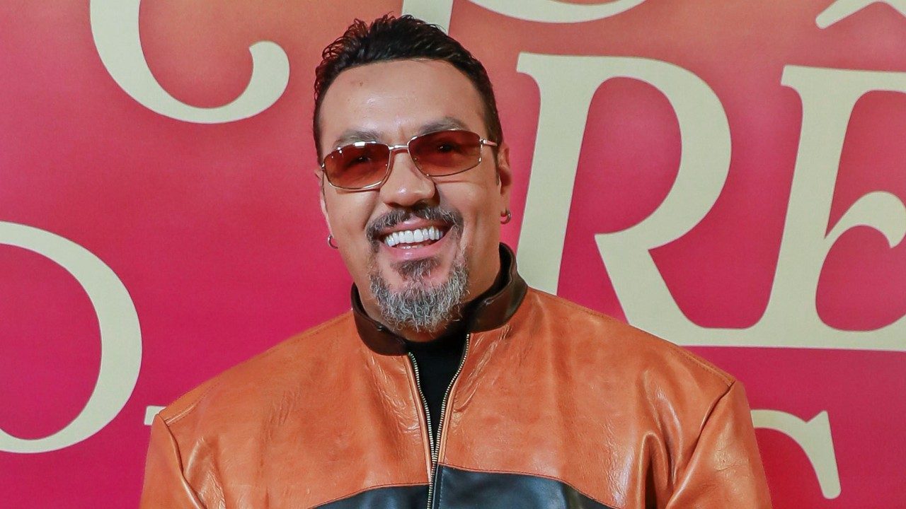 Cantor Belo sorrindo com óculos escuros e jaqueta de couro marrom diante de painel rosa em evento promocional