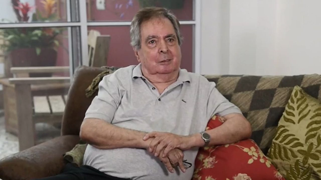 Homem idoso com camisa polo cinza fala sentado em sofá durante entrevista em casa