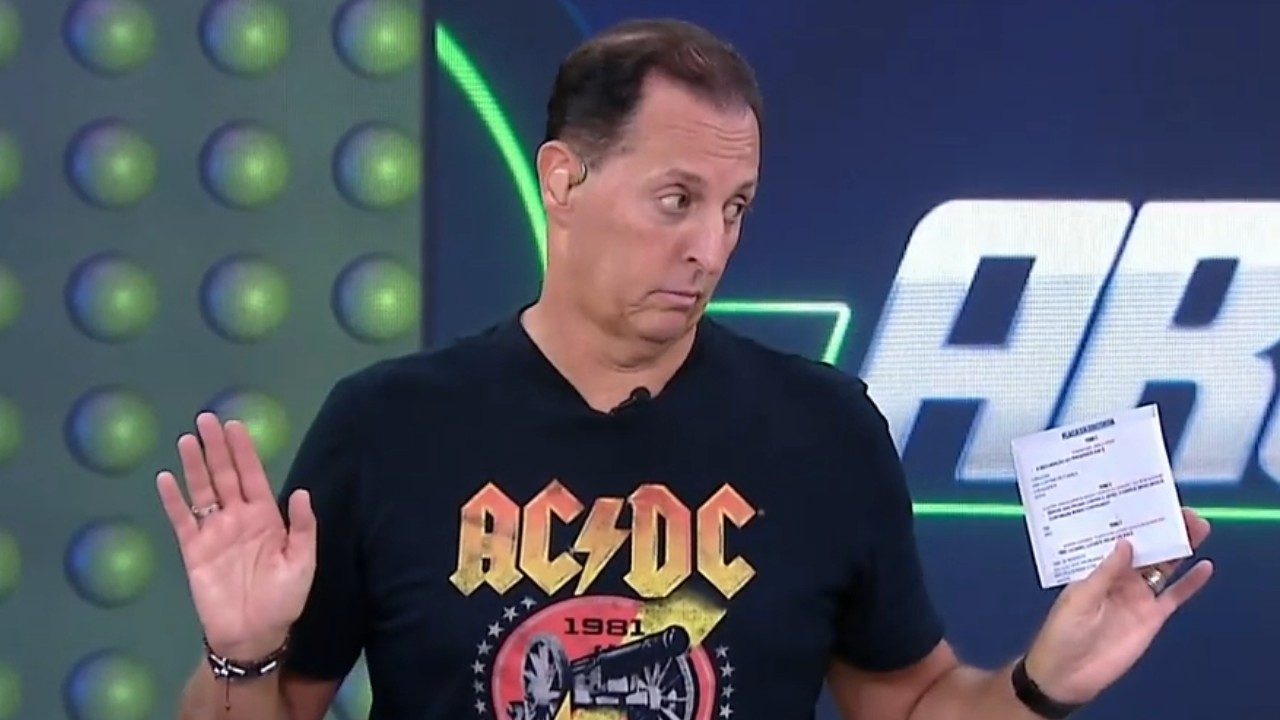 Benjamin Back sorri no cenário do programa Arena SBT, veste jaqueta jeans sobre camiseta preta, fundo colorido com luzes de neon