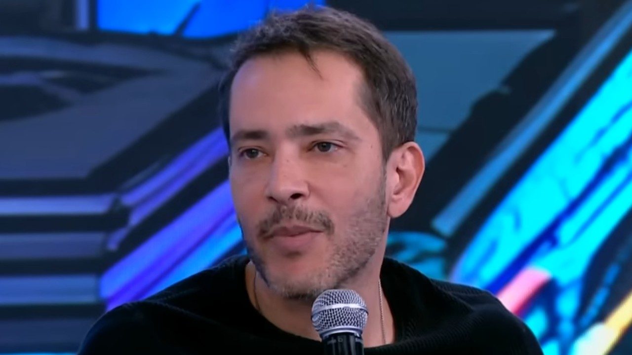Bento Ribeiro fala ao microfone durante participação em programa de televisão com cenário iluminado ao fundo