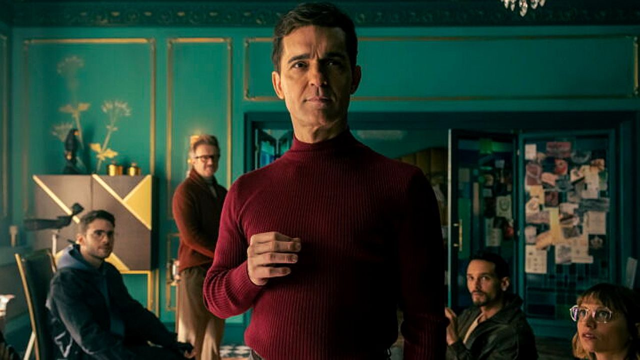 Homem de suéter vermelho fala com grupo em sala enquanto outras pessoas observam ao fundo