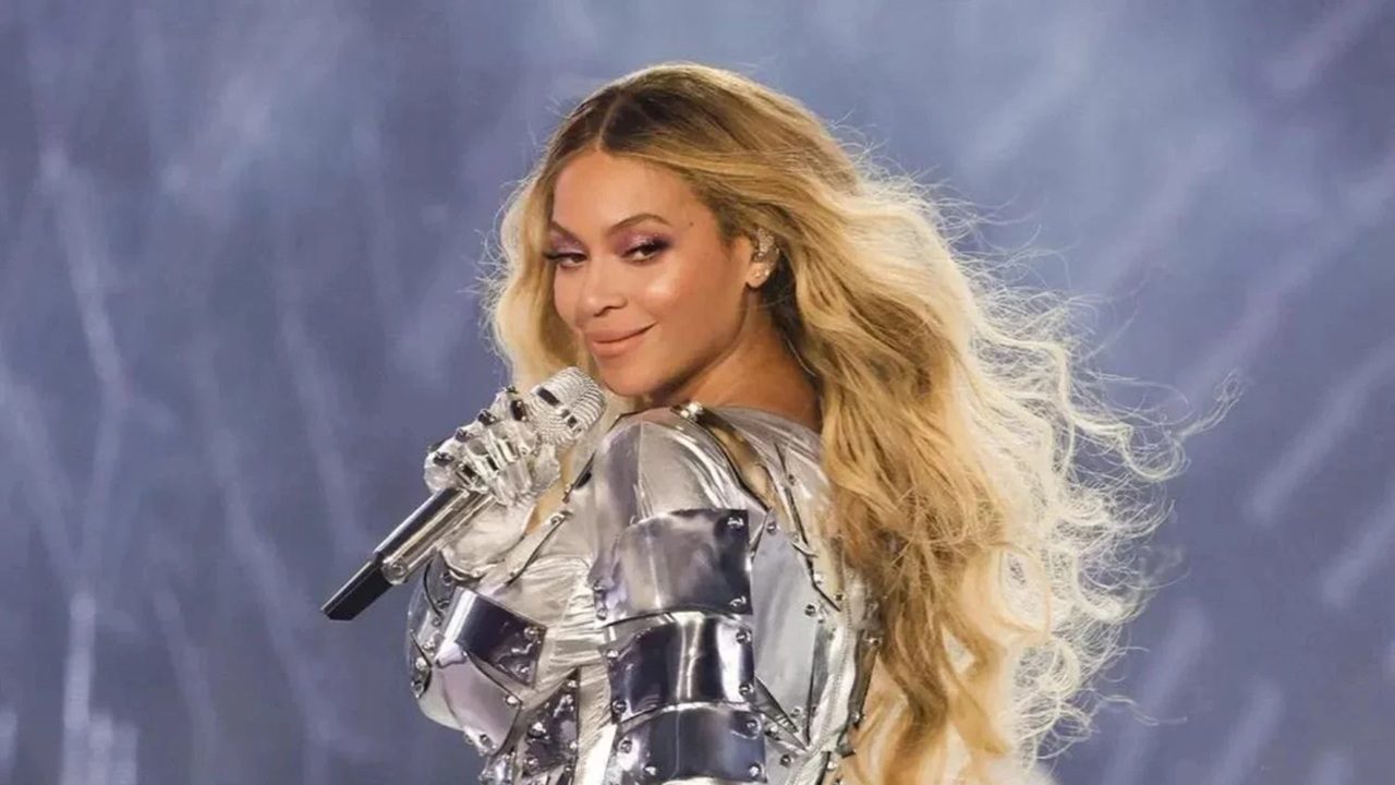 Cantora se apresentando no palco, usando figurino metálico prateado e segurando um microfone brilhante, com expressão confiante e cabelos loiros esvoaçantes sob luzes de show