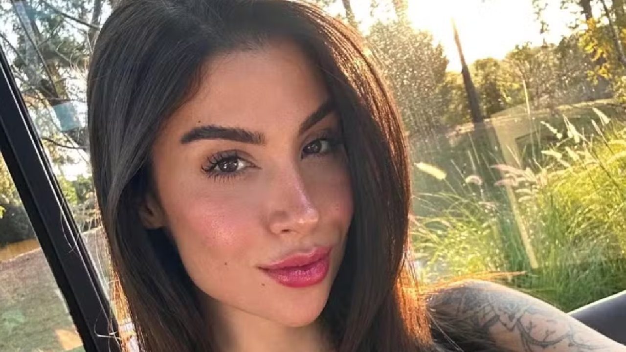 Mulher com cabelos castanhos e maquiagem leve posa ao ar livre sob a luz do pôr do sol, com vegetação ao fundo