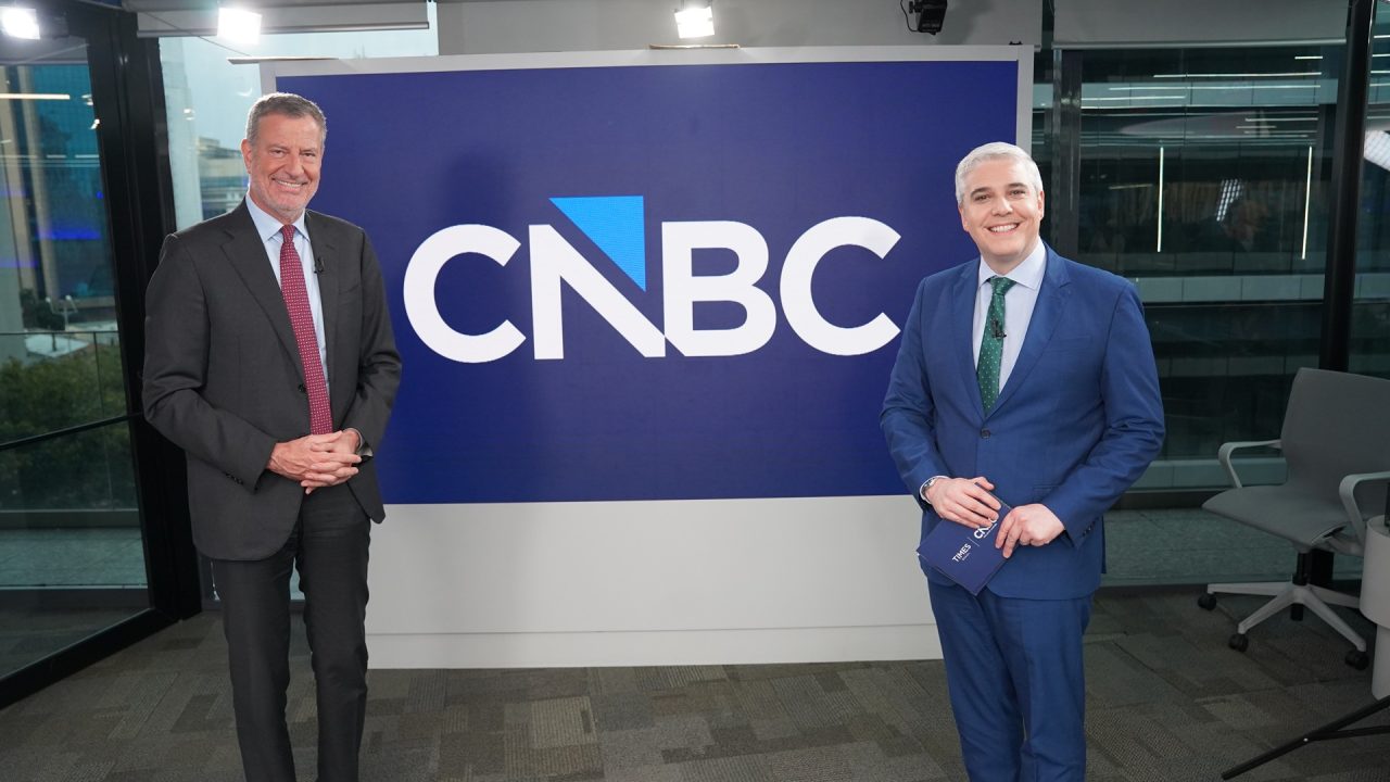 O político Bill de Blasio e o jornalista Marcelo Favalli sorriem diante de painel com logotipo da CNBC em estúdio de jornalismo econômico.