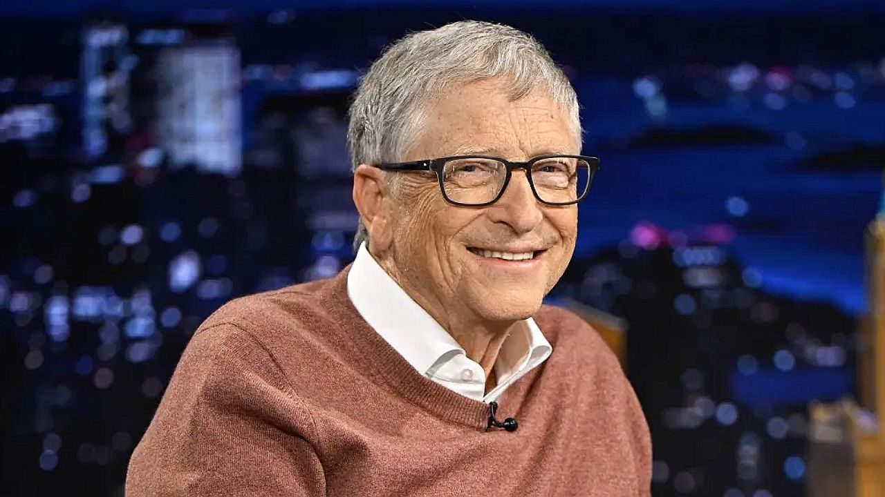 Homem idoso de cabelos grisalhos e óculos sorri durante participação em programa de televisão. Ele veste suéter marrom sobre camisa social branca, com microfone preso à roupa, diante de cenário noturno com vista urbana iluminada ao fundo.