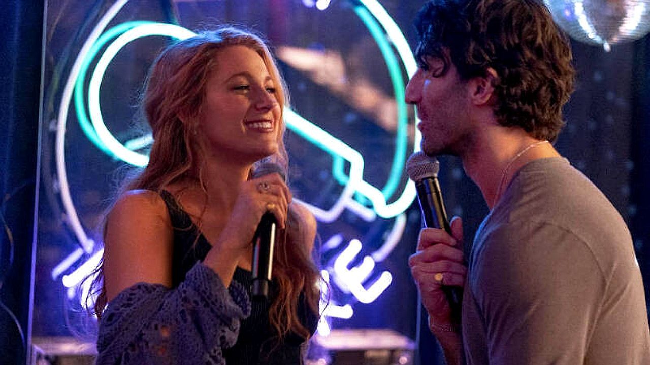 Casal canta ao microfone durante apresentação em bar com iluminação neon ao fundo em cena de filme romântico
