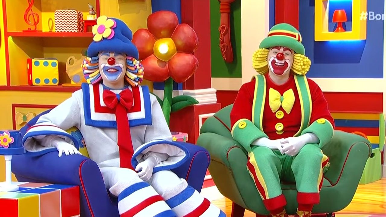 Patati (Wagner Rocha) e Patatá (Henrique Namura) sentados no cenário colorido do Bom dia e Cia no SBT
