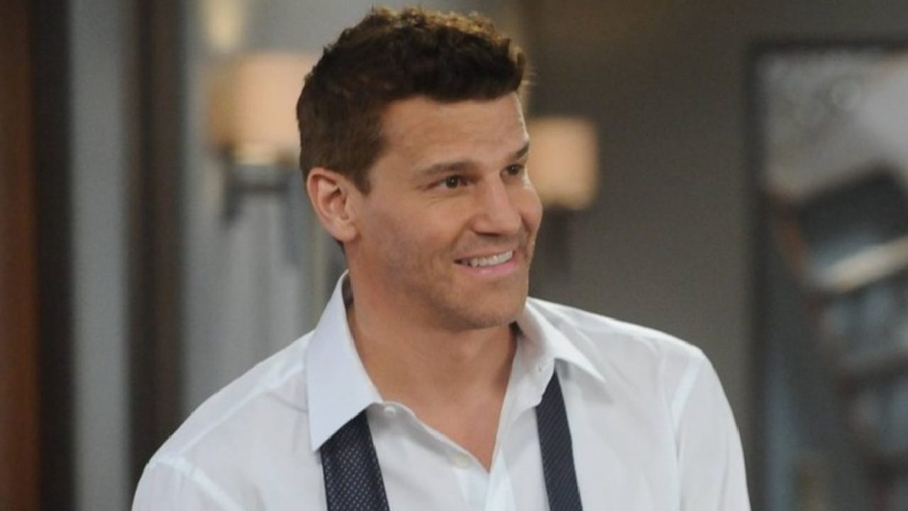 Homem de cabelo curto castanho sorri levemente enquanto olha para alguém fora de quadro. Ele veste camisa social branca com os primeiros botões abertos e suspensórios escuros, em um ambiente interno com iluminação suave e fundo desfocado.