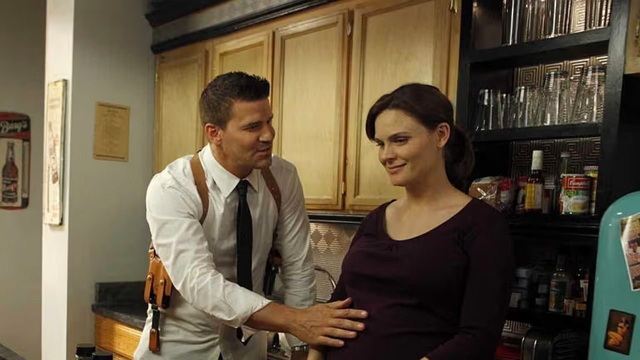 Homem de gravata e coldre toca barriga de mulher grávida em cozinha durante cena de série