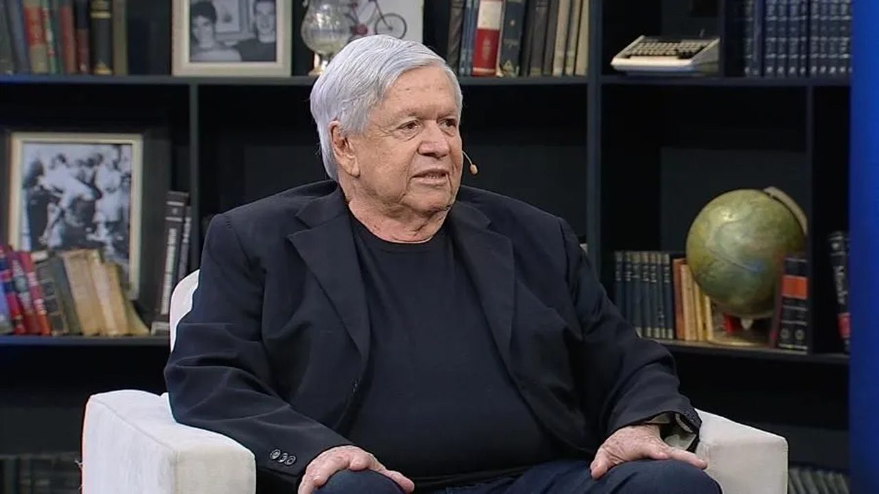 Homem idoso de cabelos grisalhos e terno escuro sentado em poltrona durante entrevista em estúdio com estante de livros e globo terrestre ao fundo