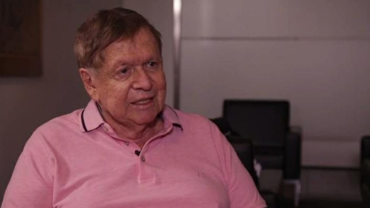 Homem de meia-idade com cabelo curto castanho claro veste camisa polo rosa e fala enquanto olha ligeiramente para o lado. Ele está em um ambiente interno com paredes claras e poltronas escuras ao fundo, sugerindo uma entrevista ou conversa em local profissional.