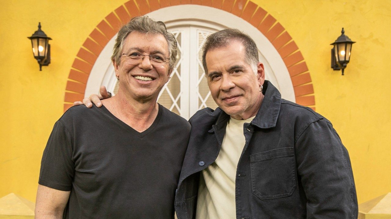 Boninho e Leandro Hassum sorriem lado a lado em frente a parede amarela com arco e luminárias em cenário de gravação