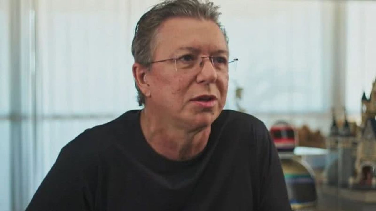 Homem de meia-idade, usando óculos e camisa preta, aparece sentado em um ambiente iluminado com objetos decorativos desfocados ao fundo. Ele olha para frente com expressão séria, como se estivesse dando uma entrevista ou explicando algo