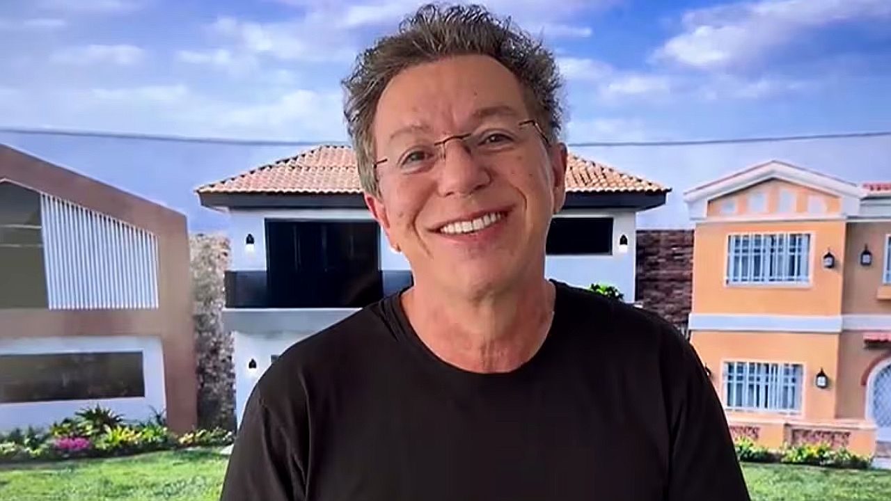 Homem sorri em frente a casas residenciais em gravação externa com céu ao fundo