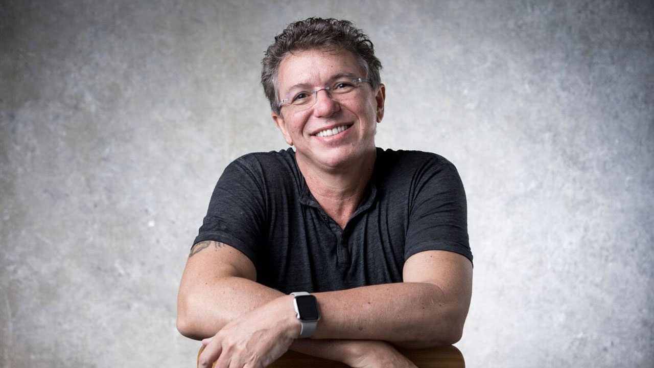 Boninho sorri apoiado em encosto de cadeira usando camisa escura e relógio branco em fundo neutro