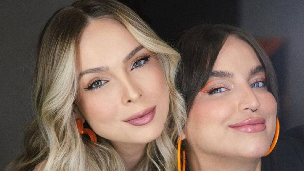 A imagem mostra duas mulheres posando juntas e sorrindo. Ambas estão maquiadas, com destaque para o delineado e os brincos laranja combinando. A mulher à esquerda tem cabelo loiro e ondulado, enquanto a da direita tem cabelo escuro e liso com franja. O fundo é neutro, destacando os rostos e os acessórios das duas