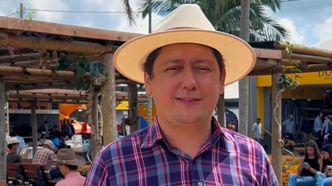 Homem de chapéu e camisa xadrez fala em área externa com barracas e pessoas ao fundo