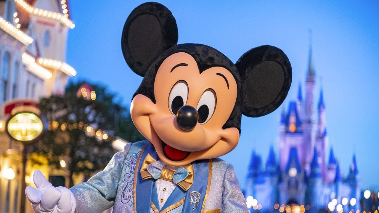 Mickey Mouse, personagem clássico da Disney, posa sorridente em traje festivo azul e dourado na Main Street do parque Magic Kingdom, com o Castelo da Cinderela iluminado ao fundo ao anoitecer.