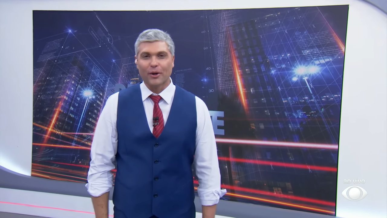 Joel Datena, vestindo um colete azul e gravata vermelha, aparece no estúdio do Brasil Urgente, com fundo digital de prédios iluminados e luzes noturnas, durante transmissão ao vivo na Band