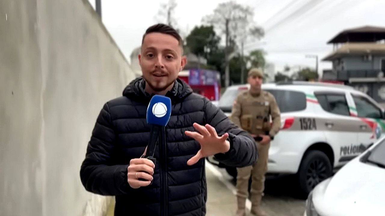 Repórter segura microfone da Record TV durante entrada ao vivo em frente a viatura policial com agente ao fundo