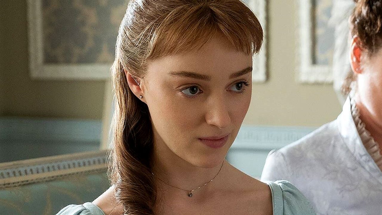 Mulher jovem com cabelos castanhos presos em penteado semi-preso aparece em ambiente interno com decoração clássica. Ela veste vestido em tom claro e colar delicado, olhando levemente para baixo com expressão atenta e contida. Ao fundo, há paredes com molduras e outra pessoa parcialmente visível, sugerindo cena de diálogo em contexto histórico ou dramático.