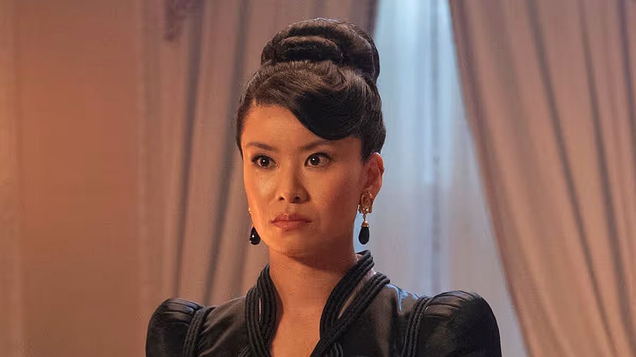 Mulher com penteado elegante e vestido preto observa à frente com expressão séria em ambiente sofisticado com cortinas ao fundo, sugerindo cena de drama ou época