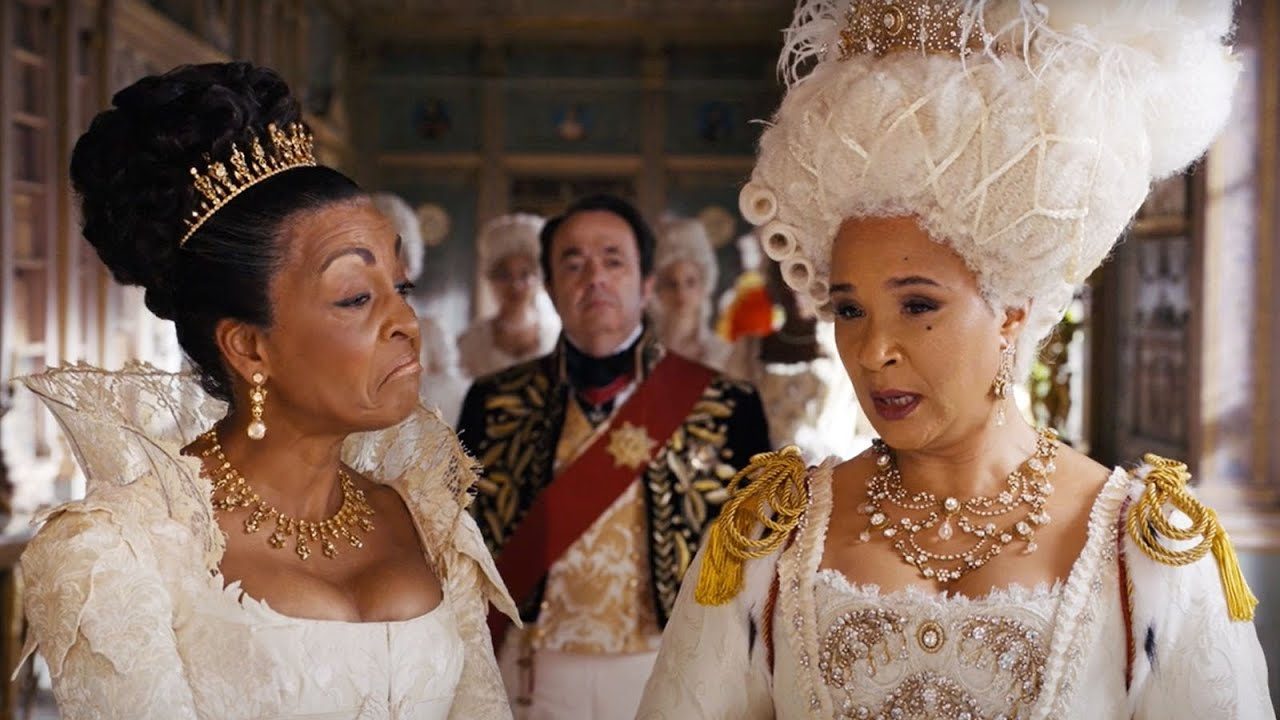 Duas mulheres em trajes de época conversam em salão luxuoso enquanto homem com uniforme nobre observa ao fundo