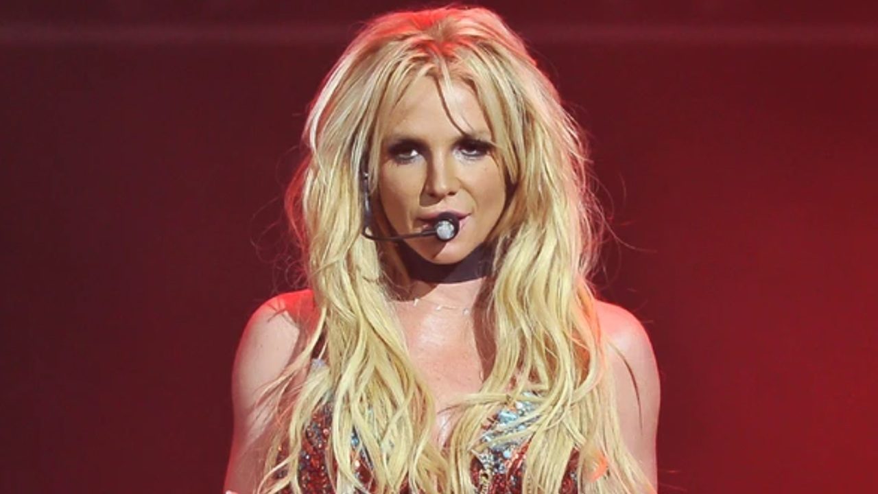 Mulher de cabelos loiros longos se apresenta no palco com microfone de cabeça e iluminação vermelha ao fundo.