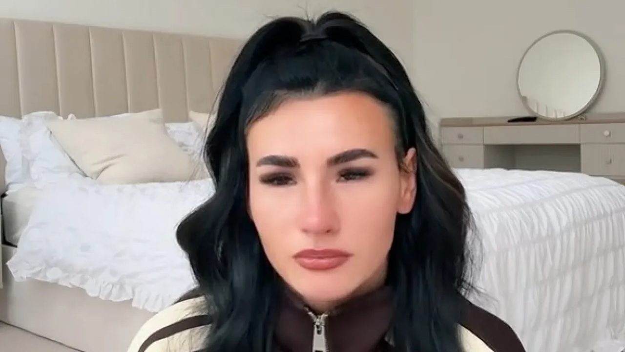 Mulher de cabelos escuros e presos no alto aparece séria em um quarto iluminado, com cama ao fundo e decoração neutra em tons claros