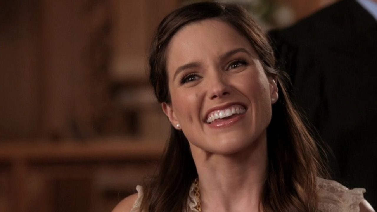 Mulher sorri amplamente em ambiente interno com iluminação quente. Ela tem cabelos castanhos penteados para trás, usa brincos discretos e uma blusa clara, transmitindo alegria e simpatia