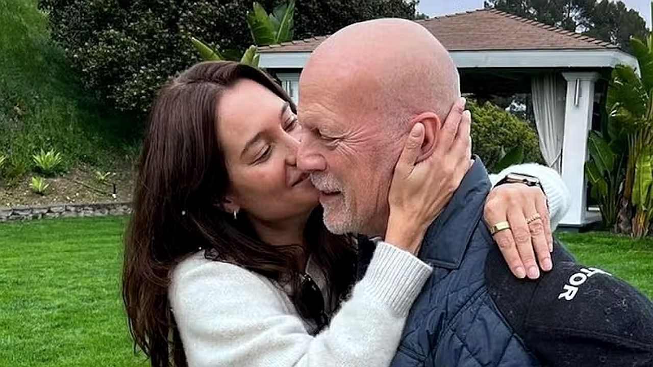 Um homem e uma mulher estão abraçados em um jardim. A mulher segura o rosto do homem enquanto o beija no rosto. Ao fundo há gramado, árvores e uma estrutura semelhante a um gazebo ou área externa de uma casa. A cena transmite um momento de carinho entre o casal.