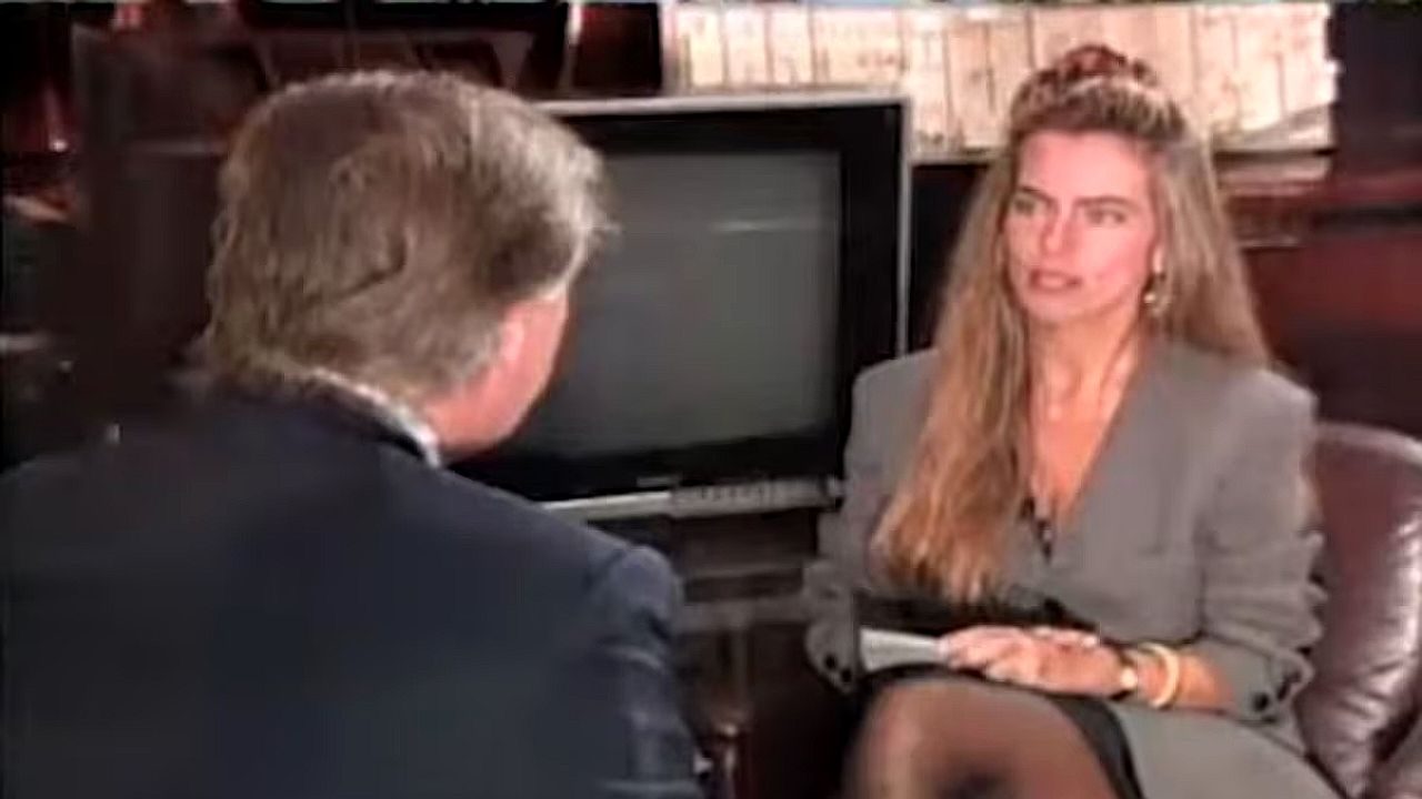 Mulher entrevista homem de terno em gravação de programa jornalístico com televisão ao fundo