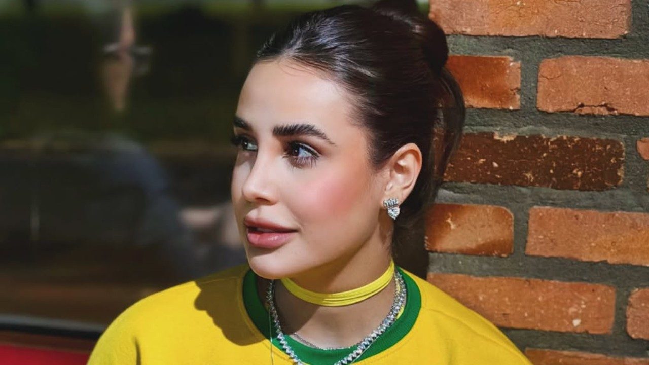 Bruna Pinheiro de blusa amarela e verde olha para o lado diante de parede de tijolos com cabelo preso e colares no pescoço