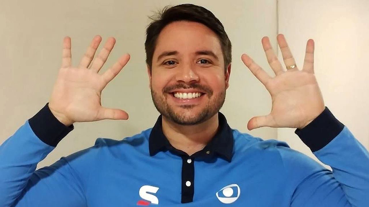 Apresentador sorri e levanta as duas mãos vestindo camisa com logotipo da Globo em fundo neutro