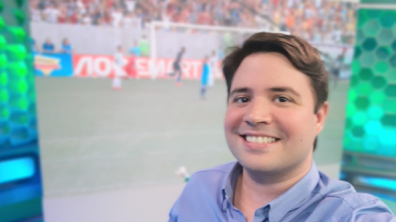 Homem sorri enquanto tira uma selfie em um estúdio de TV com temática esportiva, tendo ao fundo uma tela que exibe uma partida de futebol e arquibancada com torcedores