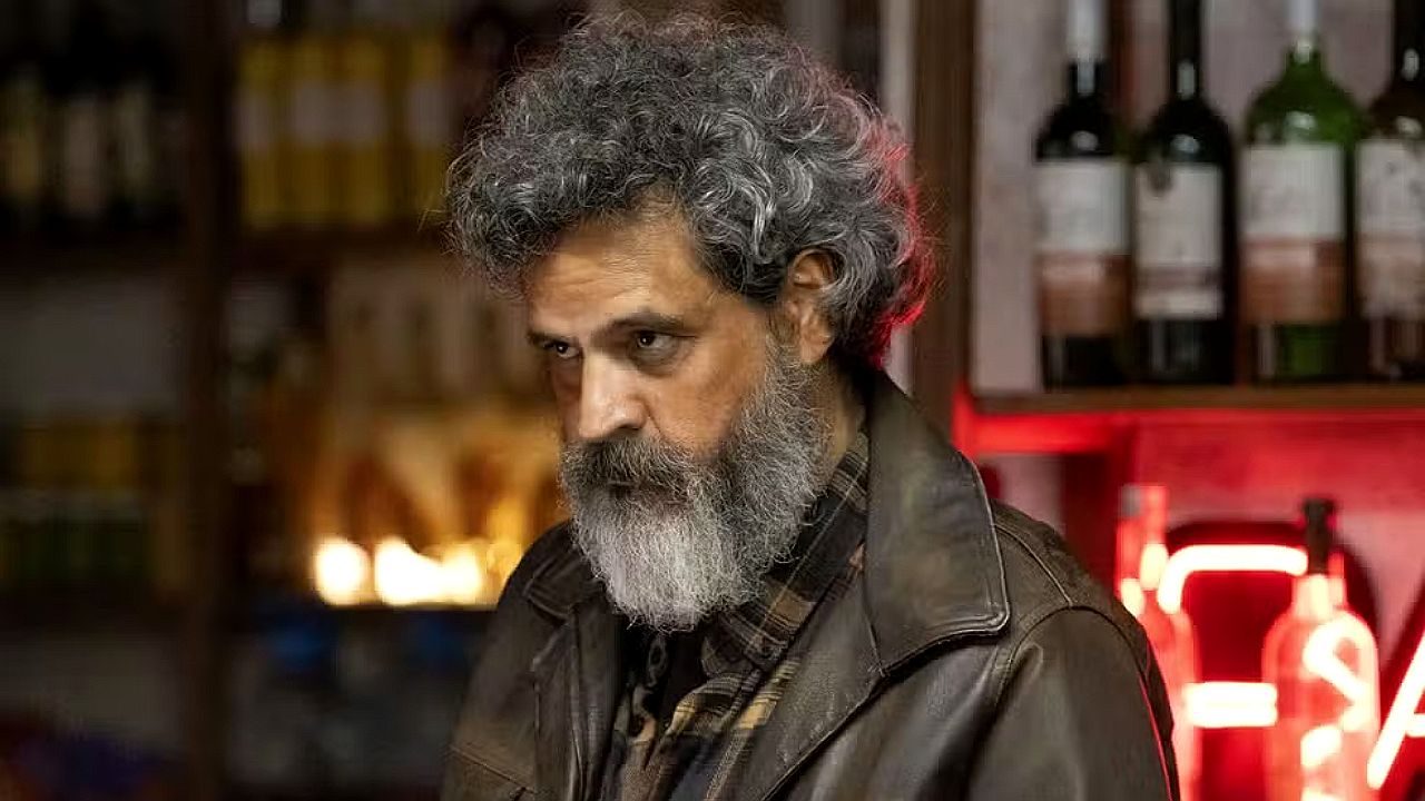 Homem de barba grisalha e casaco de couro encara sério em bar com garrafas ao fundo