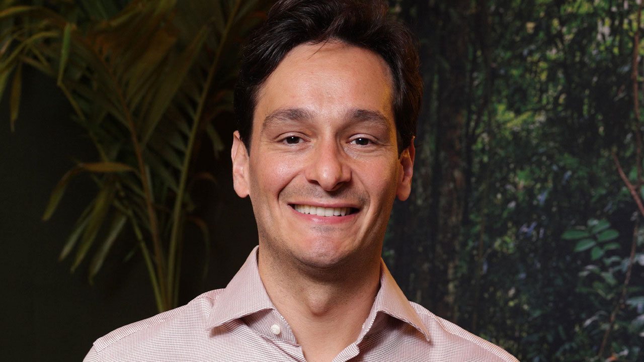 Bruno Luperi sorrindo em frente a painel com imagem de floresta usando camisa social clara