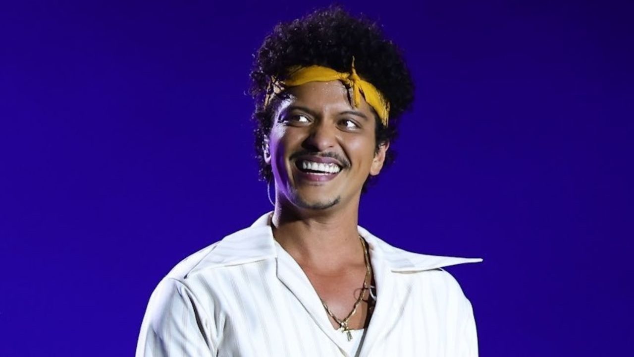 Homem sorri em frente a um fundo roxo iluminado, usando camisa branca com gola aberta, colares dourados e faixa amarela amarrada na cabeça. Ele parece estar em um palco, sob luzes de show.